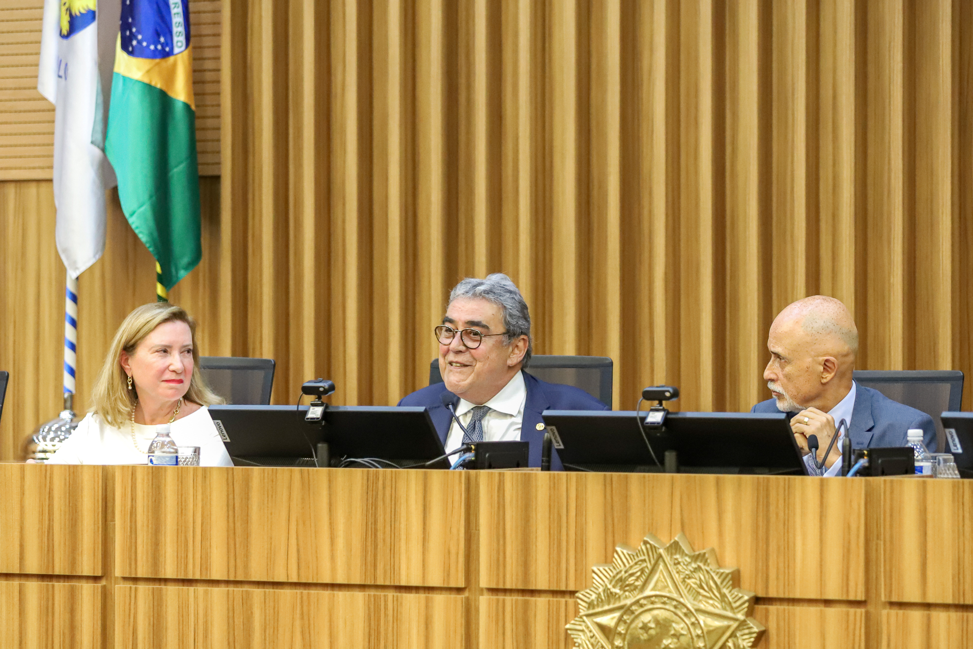 ministro Aloysio Veiga fala ao microfone, sendo observado pelos desembargadores Rita Penkal, à sua esquerda, e Samuel Lima, à direita