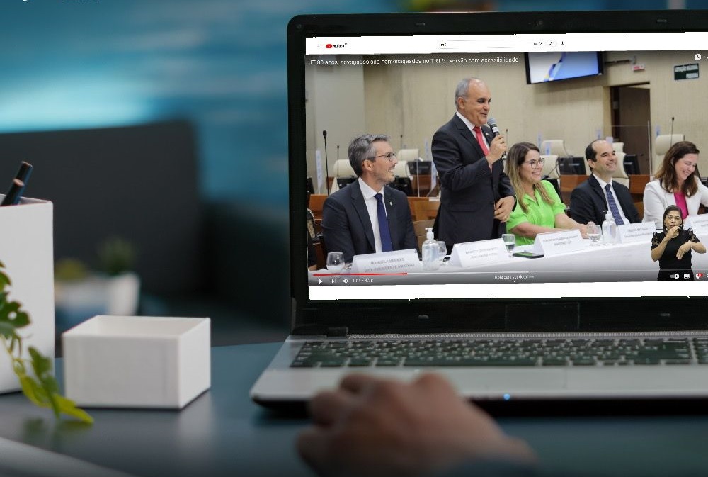 Imagem de um notebook ligado sobre uma mesa exibindo um dos vídeos com audiodescrição e janela de Libras do Canal do TRT-5 no YouTube