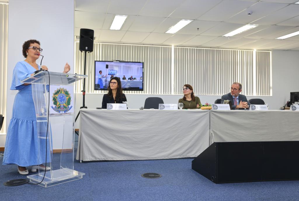 mulher fala ao microfone e autoridades estão sentadas à mesa